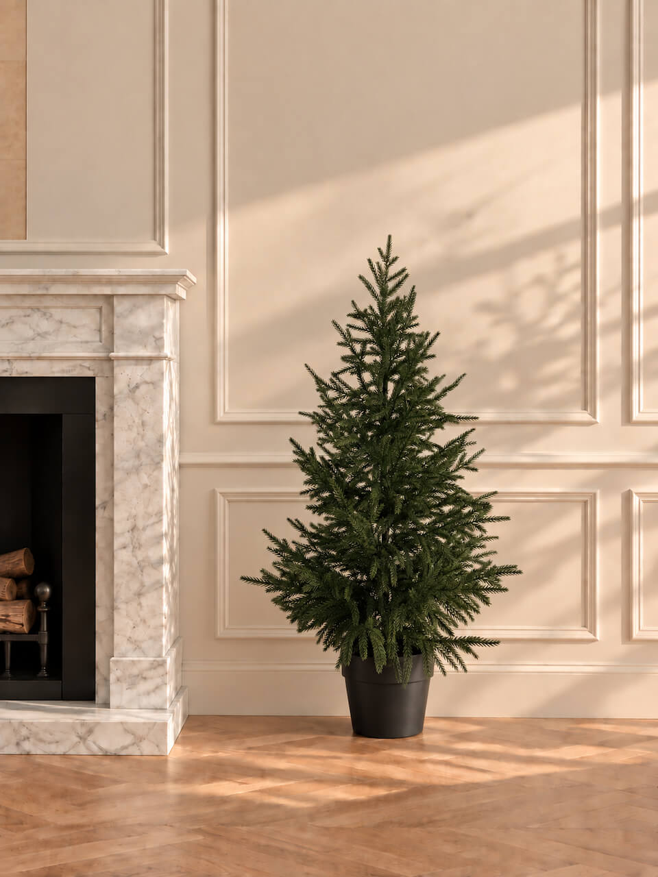 A dense artificial Christmas tree in a black pot, placed on a wooden floor next to a white marble fireplace in a room with beige paneled walls and warm sunlight.