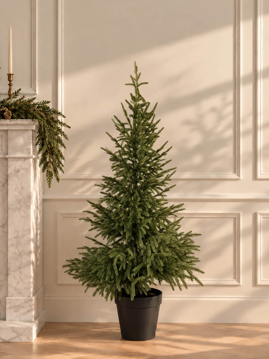 A tall, green artificial Christmas tree in a black nursery pot, set against a cream-colored paneled wall with warm sunlight casting soft shadows.
