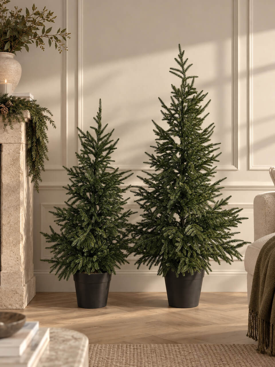 Two artificial Christmas trees in black pots, one smaller and one larger, displayed in a cozy living room setting with a fireplace and wood flooring.