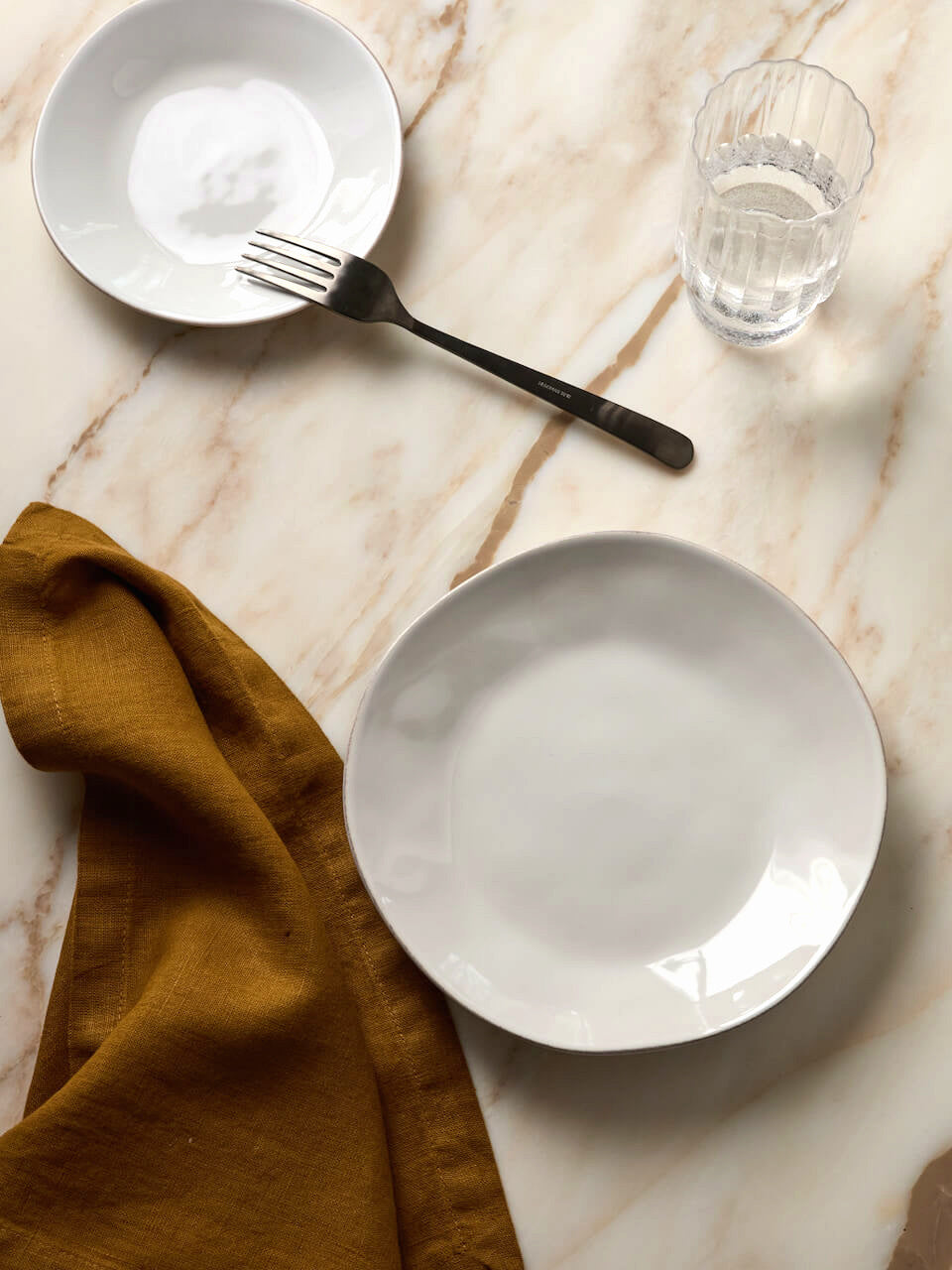Two white plates, a black fork, and a glass of water on a marble surface with a mustard-colored napkin.