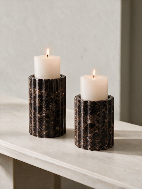 Two dark brown marble candle holders with a fluted cylindrical design, each holding a lit white pillar candle, set on a light stone surface against a neutral textured wall.