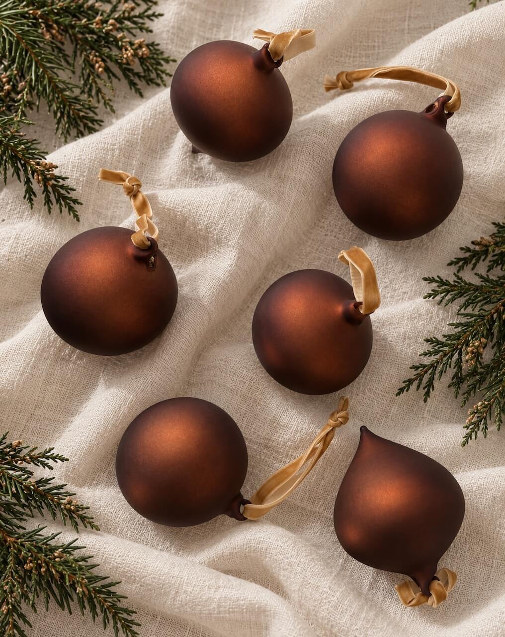 Six matte dark brown Christmas ornaments in various shapes, including three round balls and three teardrop ornaments, each with a light tan ribbon hanger, arranged on a textured cream linen fabric with green pine branches in the corners.