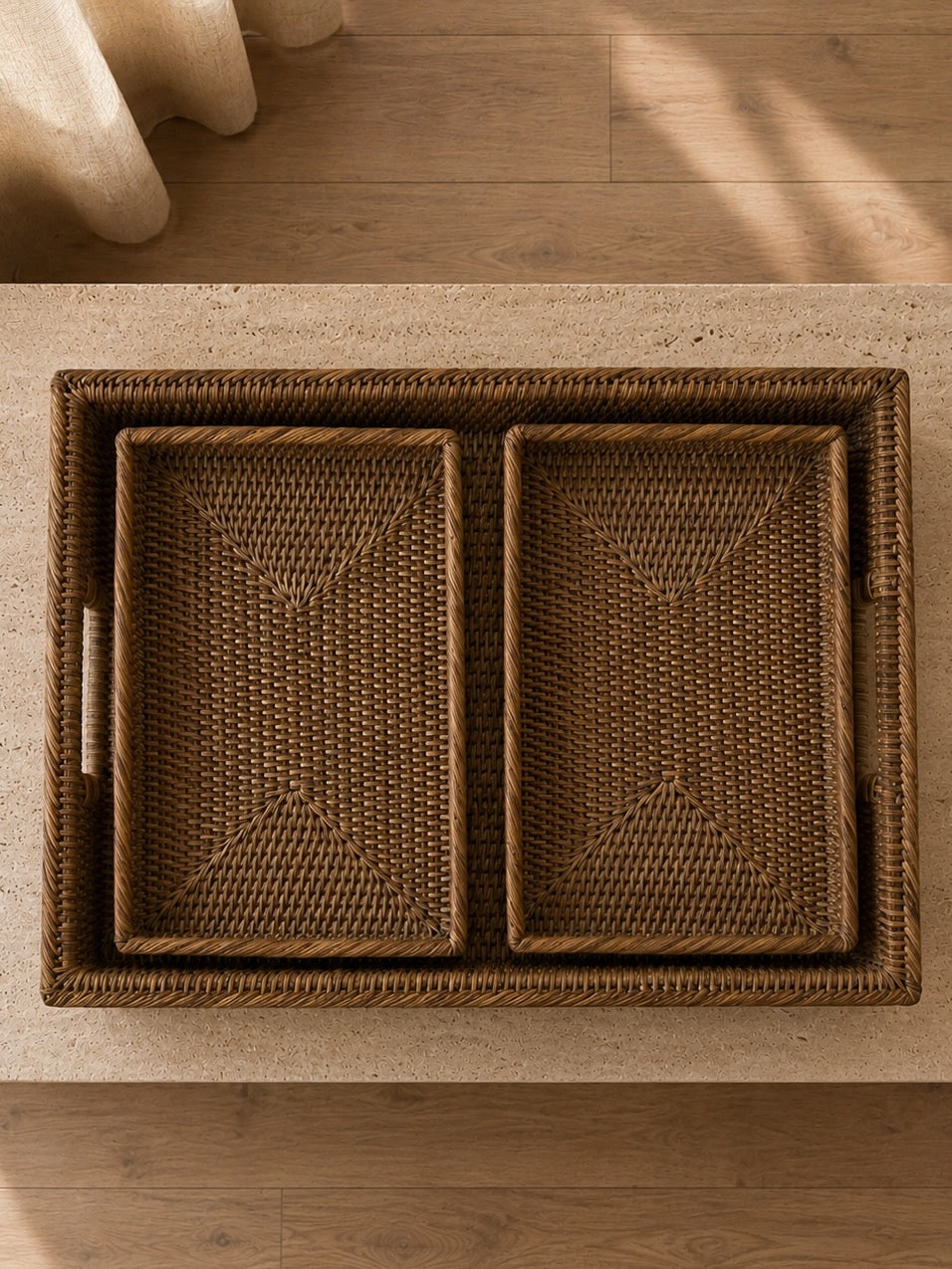 Overhead view of a handwoven antique brown rattan tray set with two smaller trays nested inside, styled on a travertine coffee table with warm wooden flooring below.