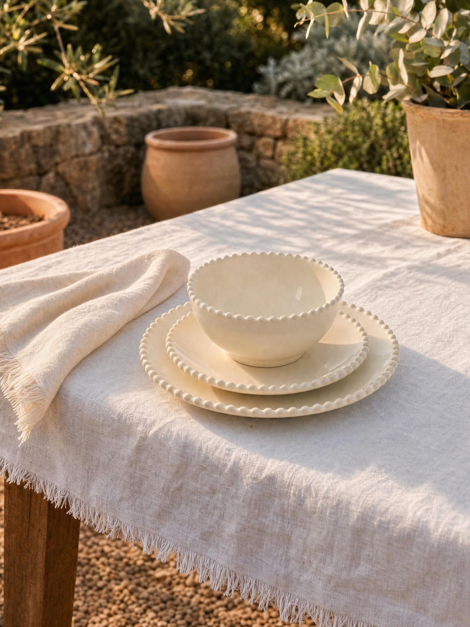 Cream scalloped dinnerware set with cereal bowl, side plate, and dinner plate stacked on a white linen table outdoors in a warm Mediterranean garden setting.