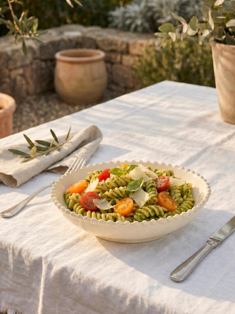 Cream scalloped pasta bowl filled with pesto fusilli, cherry tomatoes, basil, and shaved parmesan, styled on a white linen table outdoors in a sunlit Mediterranean garden setting.