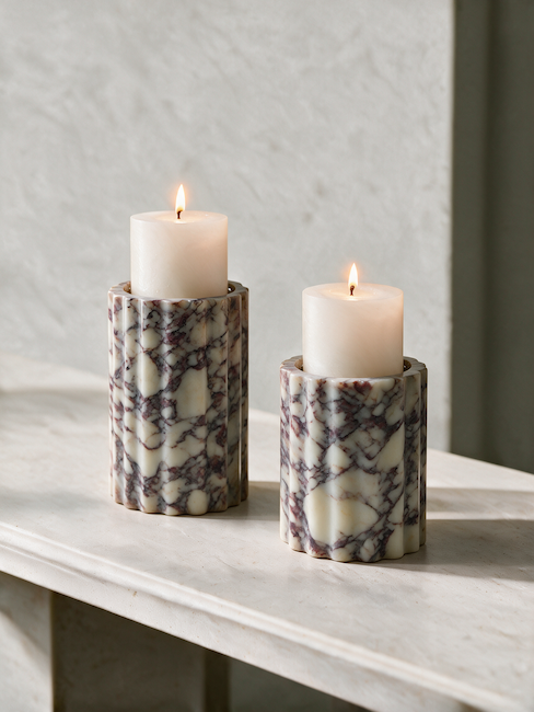 Two white pillar candles in ribbed cylindrical marble holders with a cream and purple marble Calcutta Viola pattern, set on a light stone surface against a textured light gray wall.