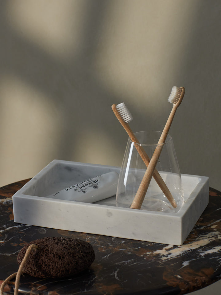 A rectangular Carrara marble tray with a soft grey vein, displayed on a bathroom counter with toothbrushes and a glass cup.