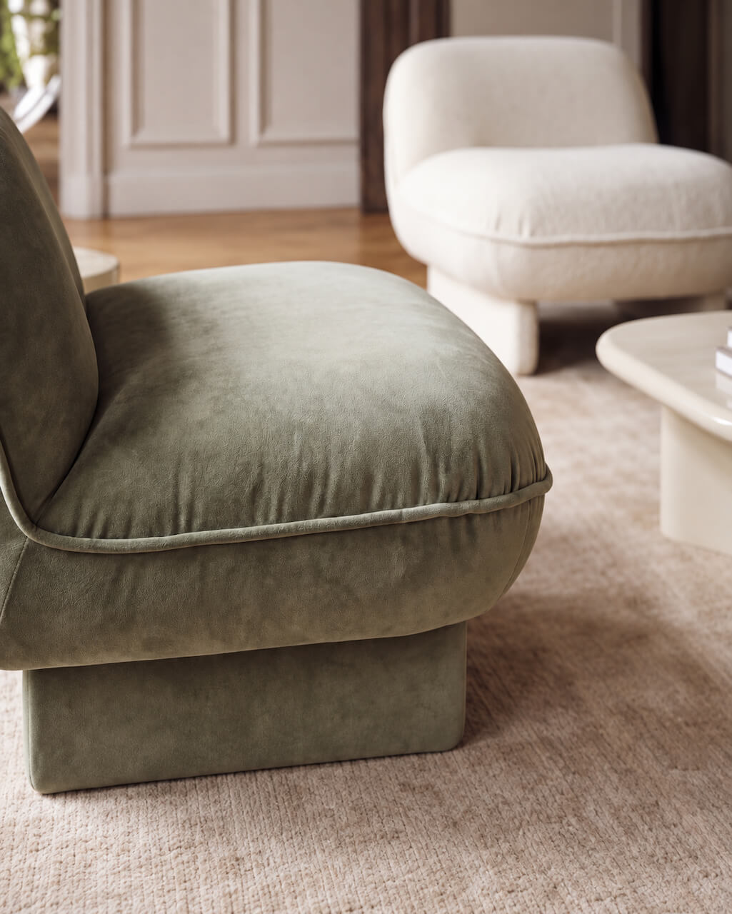 A close-up of a modern upholstered armchair in muted sage green velvet, set on a textured beige rug in a softly lit living room with another chair and a coffee table in the background.