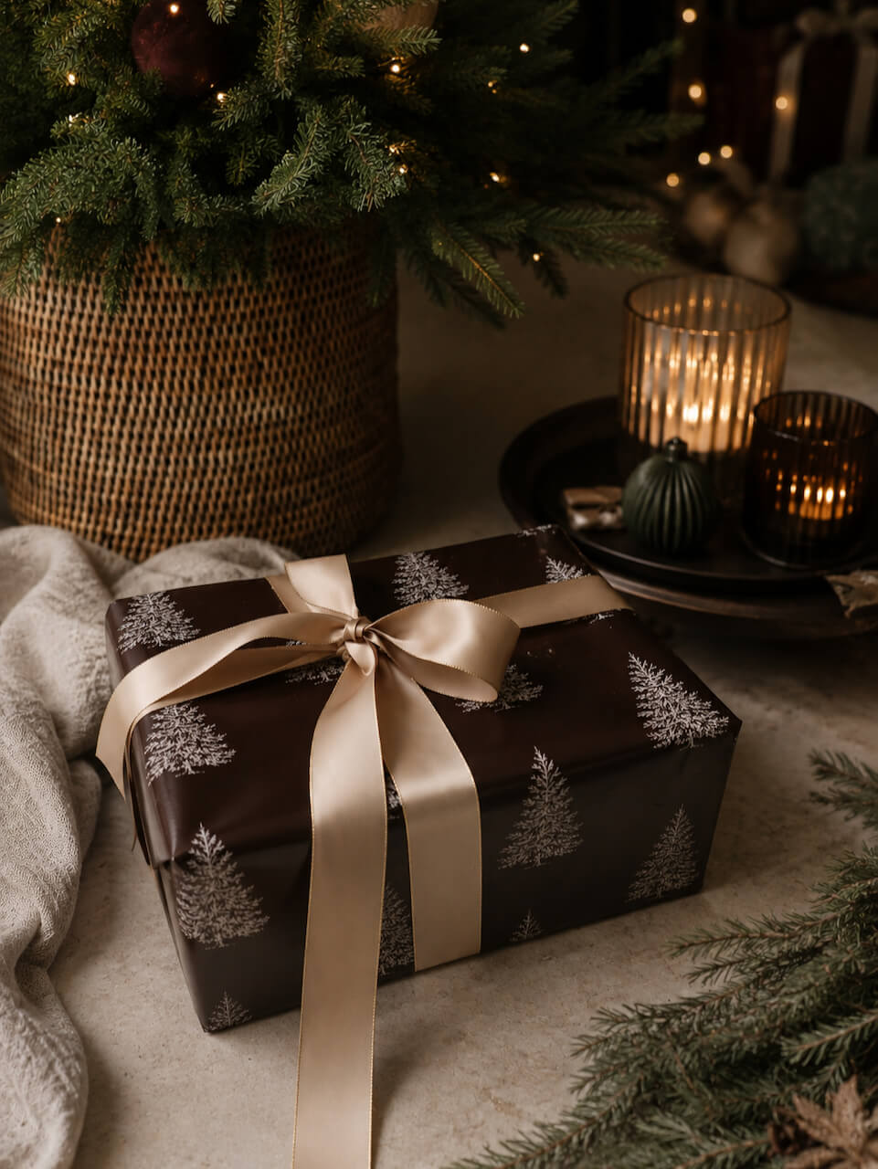 A wrapped gift box with dark brown paper featuring a white pine tree pattern, tied with a wide champagne-gold satin ribbon, set on a neutral surface with holiday decor including a wicker basket, candles, and evergreen branches.