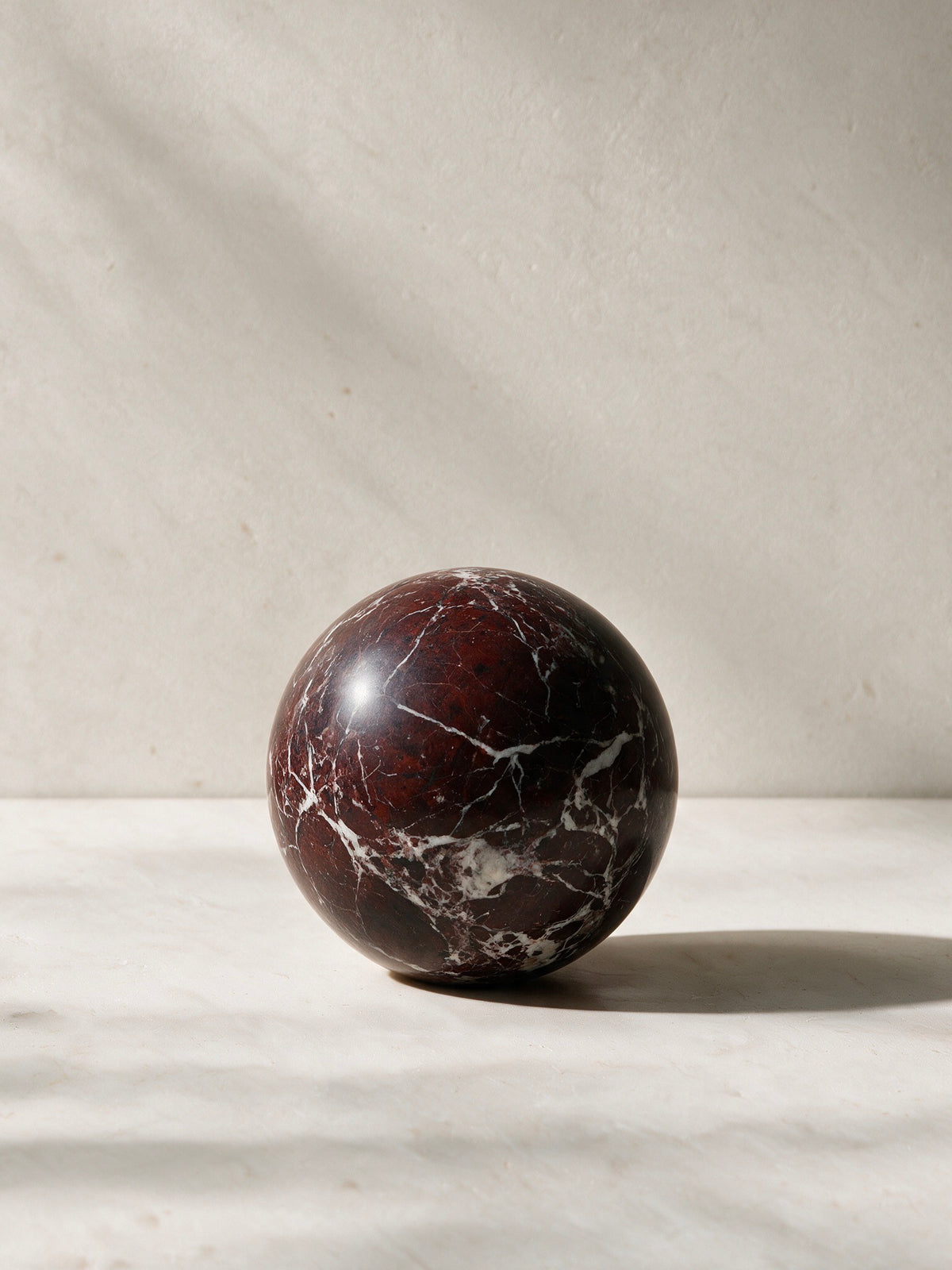 A polished spherical stone sculpture in a deep reddish-brown marble pattern with white veining, set against a neutral textured beige background.