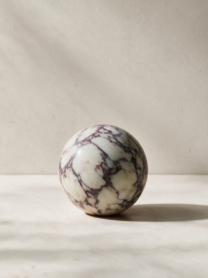A polished spherical stone sculpture with a white base and dark brown to black veining, displayed on a light marble surface against a neutral beige background.