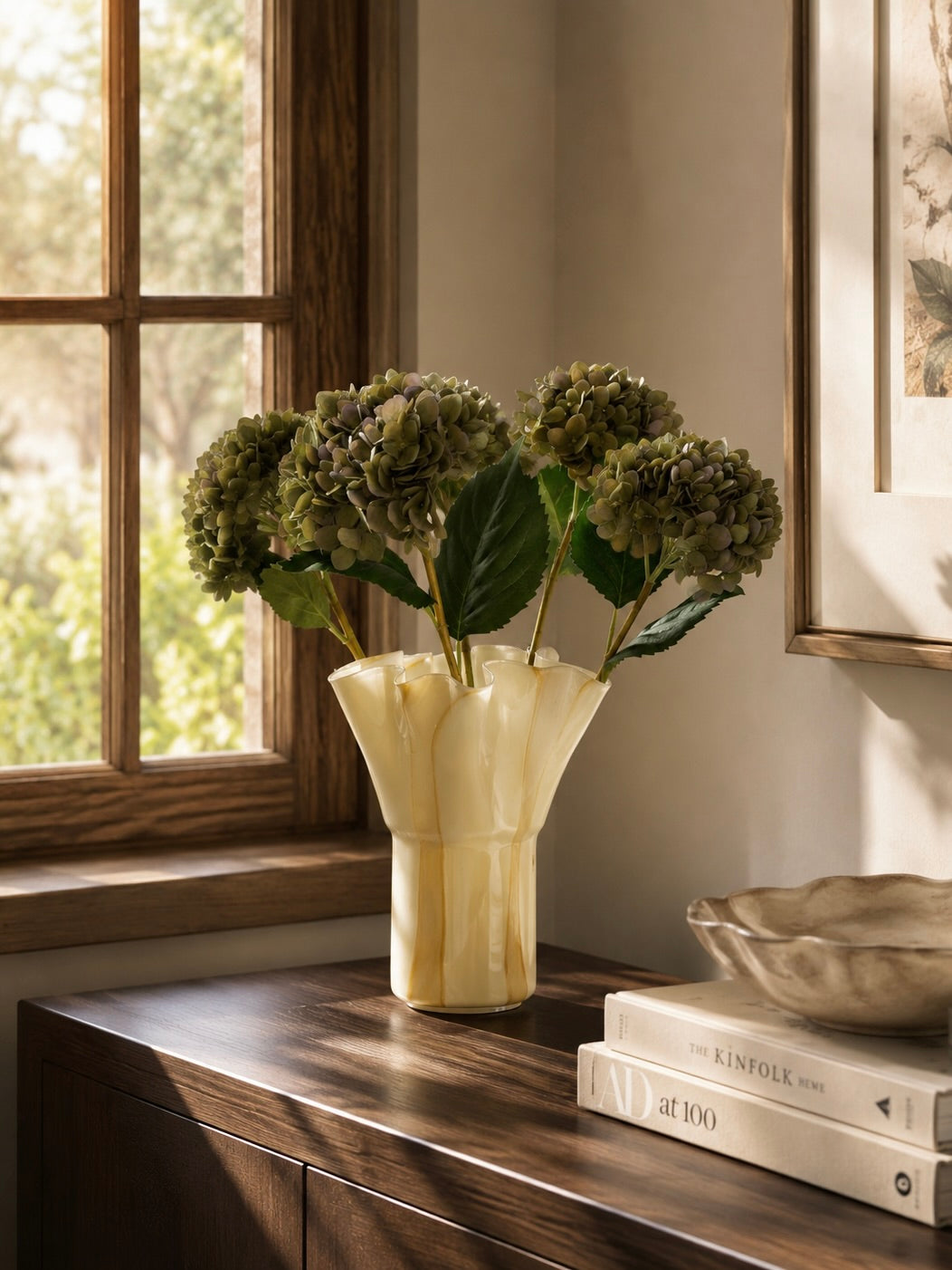 Vintage green faux hydrangea stems arranged in a cream marbled ruffle vase on a dark wood sideboard beside a wooden-framed window, styled in a warm neutral living room with natural sunlight.