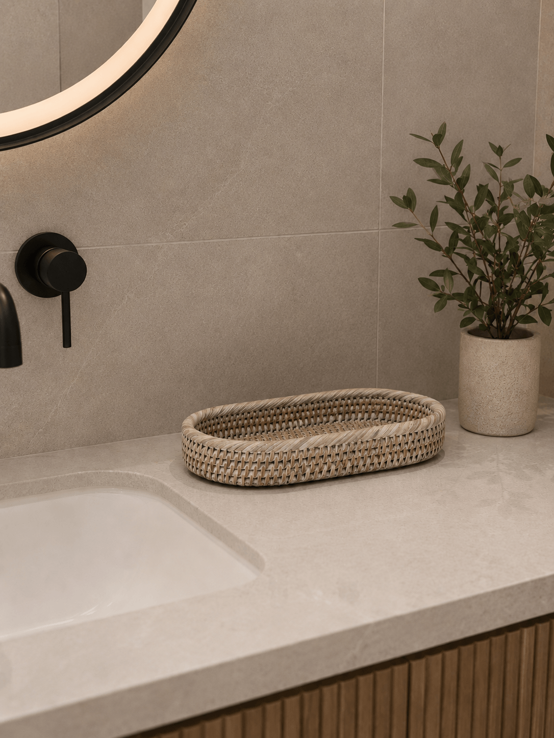 Empty white rattan bathroom tray displayed on a light stone countertop beside a potted plant and matte black bathroom fixtures in a modern neutral bathroom.