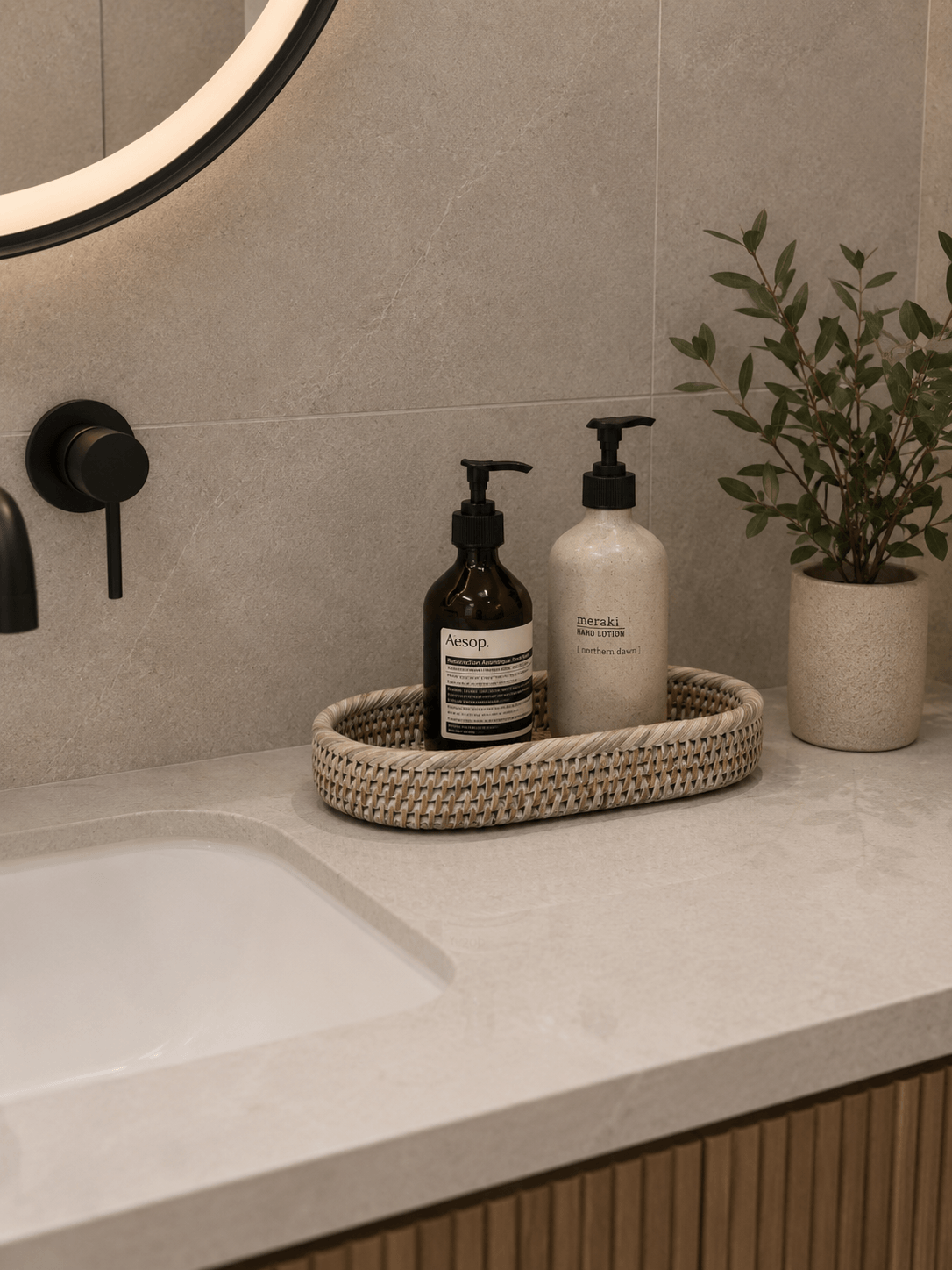 Oval white rattan bathroom tray holding amber and speckled lotion bottles on a beige stone vanity with black fixtures and a decorative plant in the background.