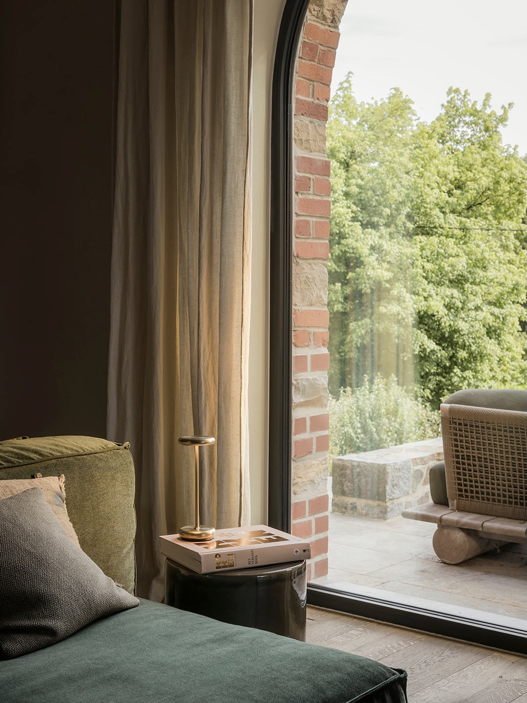 A styled interior corner showing a dark green velvet sofa with a gray pillow, a small side table with a brass table lamp and a book, and a large arched window looking out onto a brick patio and lush green trees.