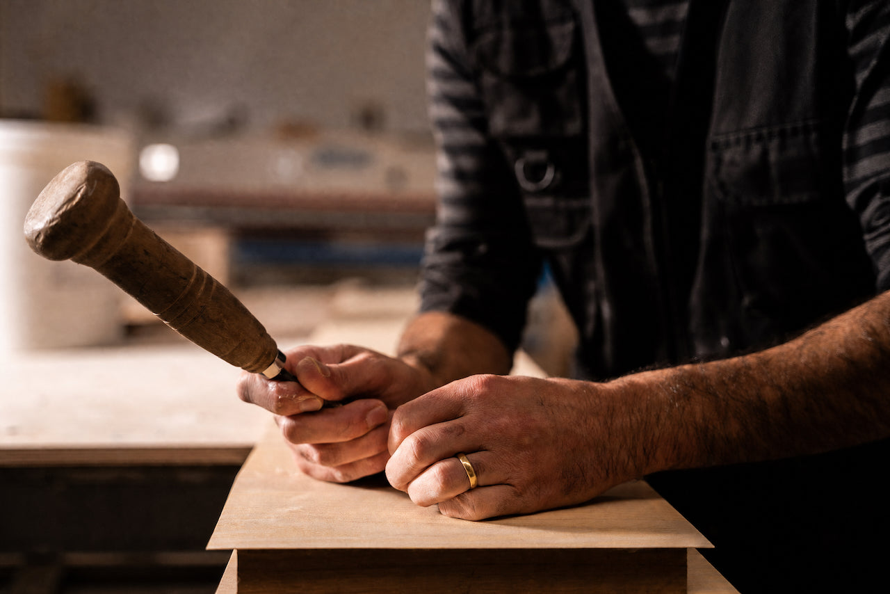 A close-up lifestyle shot of a craftsman's hands holding a wooden mallet and chisel, carving or marking a piece of light-colored wood in a workshop setting.