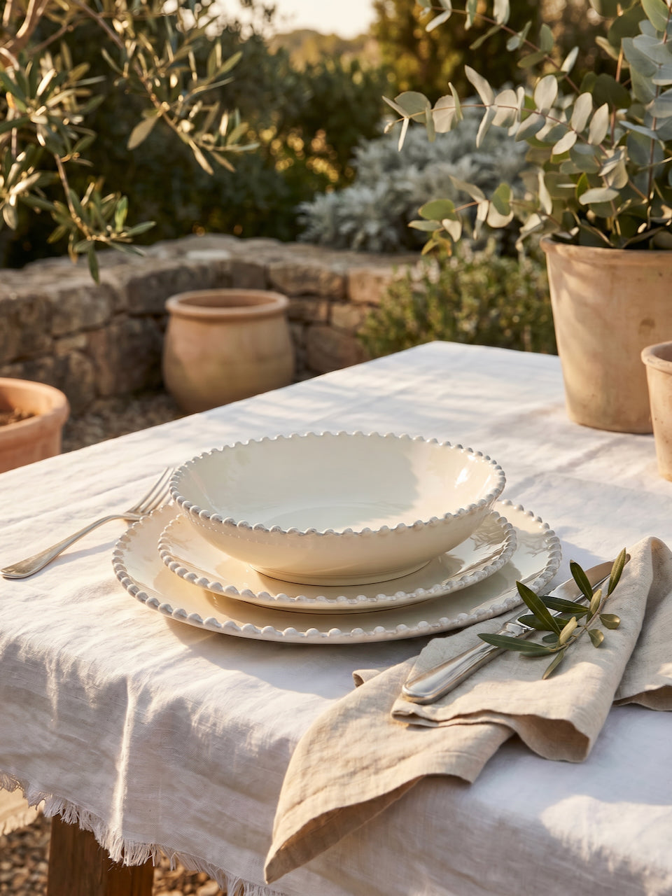 Cream scalloped dinnerware set including dinner plate, side plate, and pasta bowl styled on a white linen table outdoors with olive branches and Mediterranean garden surroundings.