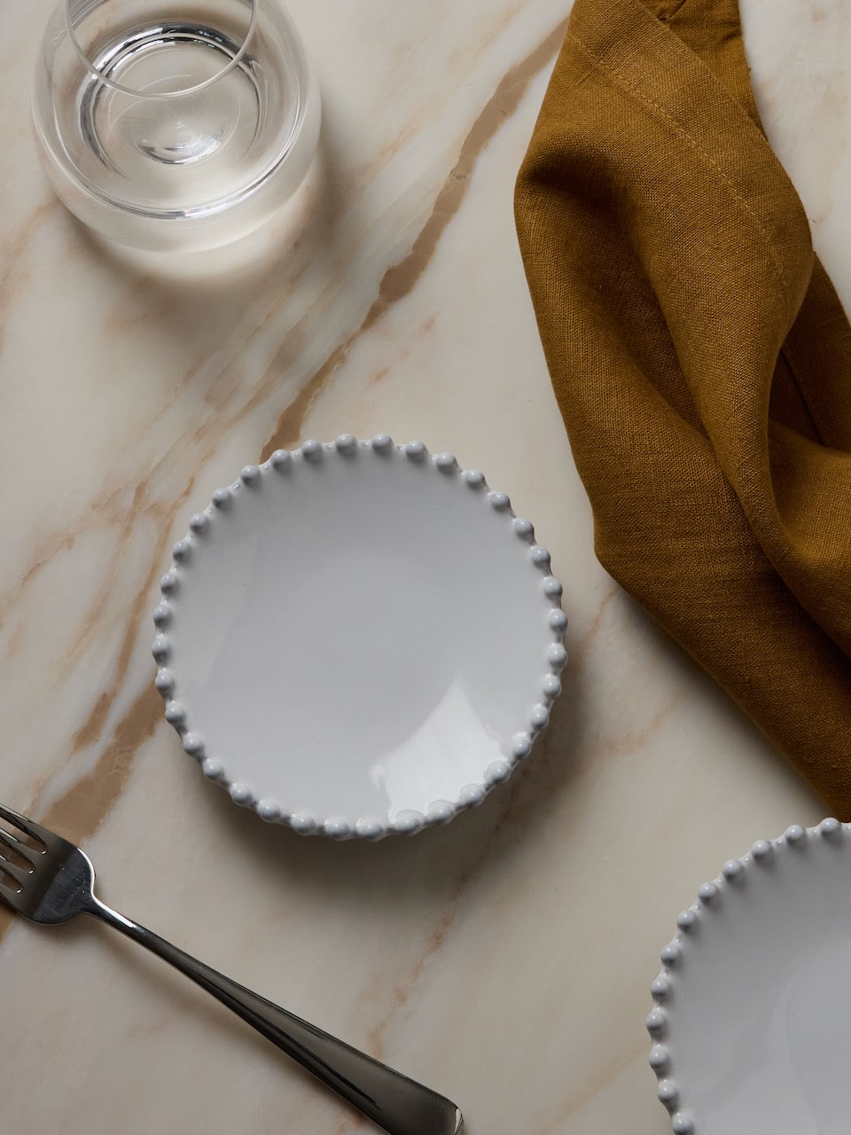 White ceramic plate with scalloped edge on a marble surface with a fork and bronze coloured napkin.
