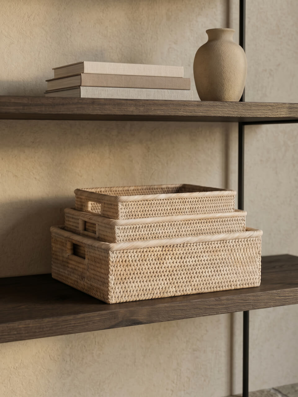Set of three woven natural rattan storage trays stacked on dark wooden shelves with neutral styling, ceramic vase and linen books in a warm minimalist interior.