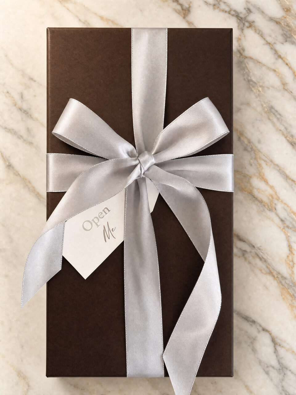 A dark brown rectangular gift box wrapped with a wide silver satin ribbon and bow, with a small white tag reading 'Open Me,' placed on a light marble surface.