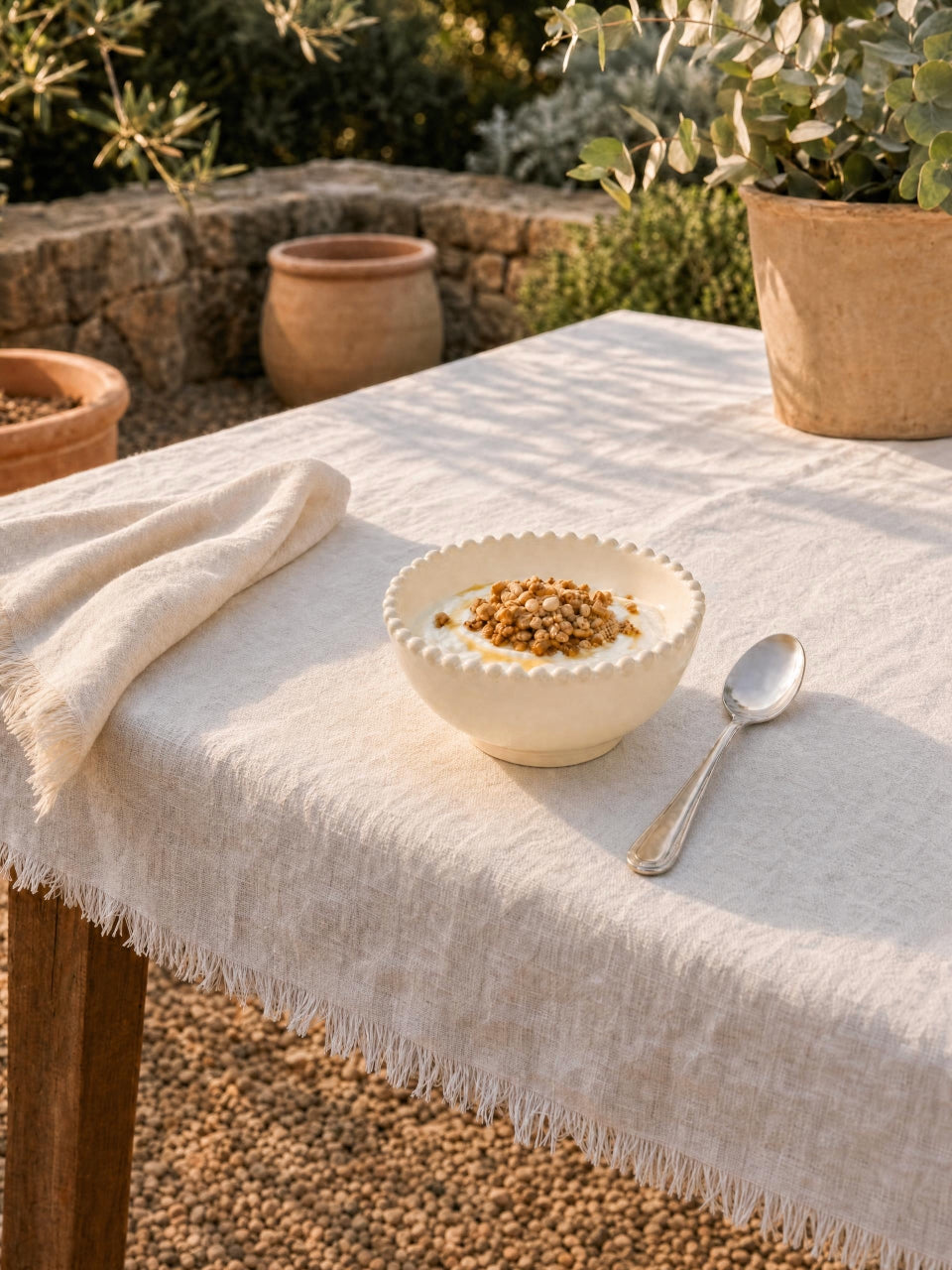 Cream scalloped cereal bowl filled with yoghurt and granola, styled on a white linen table outdoors with warm Mediterranean garden surroundings and soft evening light.